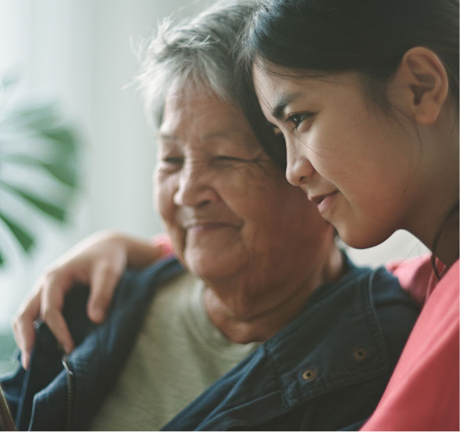 An image of a two women in a side hug. One woman looks like an eldery woman and a younger woman is giving her a side hug and they are both smiling